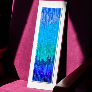 May include: A rectangular artwork with a gradient of blue and teal glass rods set in a white frame. The artwork is leaning against a magenta upholstered chair, with a wooden arm visible.