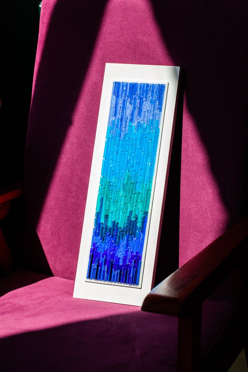 May include: A rectangular artwork with a gradient of blue and teal glass rods set in a white frame. The artwork is leaning against a magenta upholstered chair, with a wooden arm visible.