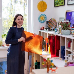 K&ouml;nnte beinhalten: Eine Frau in einem blauen Hemd und einer Sch&uuml;rze h&auml;lt ein gro&szlig;es St&uuml;ck orangefarbenes Glas in einer Werkstatt. Die Werkstatt ist gef&uuml;llt mit Regalen voller bunter Glasscheiben und anderer Kunstmaterialien.