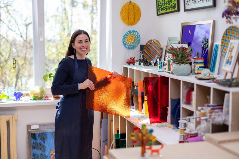 K&ouml;nnte beinhalten: Eine Frau in einem blauen Hemd und einer Sch&uuml;rze h&auml;lt ein gro&szlig;es St&uuml;ck orangefarbenes Glas in einer Werkstatt. Die Werkstatt ist gef&uuml;llt mit Regalen voller bunter Glasscheiben und anderer Kunstmaterialien.