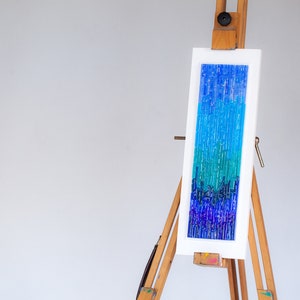 May include: Abstract art piece with a vertical rectangular design in shades of blue and green. The artwork is displayed on a white background within a white frame, mounted on a wooden easel. The piece evokes a sense of depth and texture.