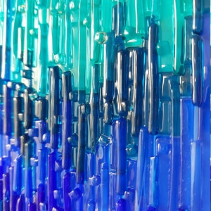 May include: Abstract art piece featuring vertical glass rods in shades of blue and teal. The rods vary in length and thickness, creating a textured, layered effect. The color gradient transitions from light teal at the top to deep blue at the bottom.
