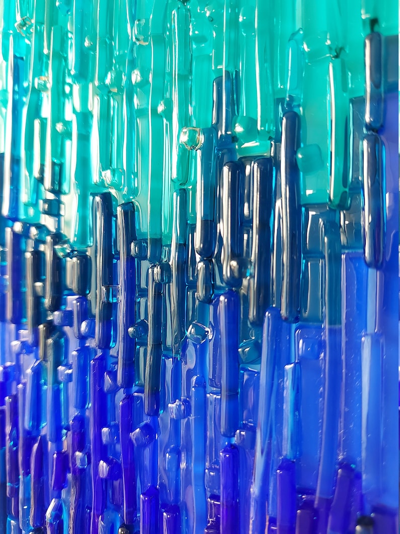 May include: Abstract art piece featuring vertical glass rods in shades of blue and teal. The rods vary in length and thickness, creating a textured, layered effect. The color gradient transitions from light teal at the top to deep blue at the bottom.