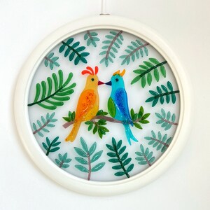 May include: A circular glass art piece with a white frame, featuring two colorful birds perched on a branch surrounded by green leaves.