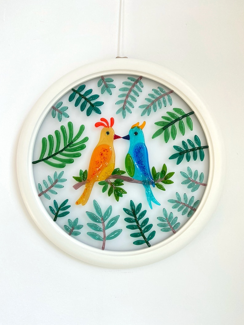 May include: A circular glass art piece with a white frame, featuring two colorful birds perched on a branch surrounded by green leaves.