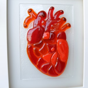 May include: A framed glass mosaic artwork depicting a red heart. The heart is made of many small pieces of red and orange glass.