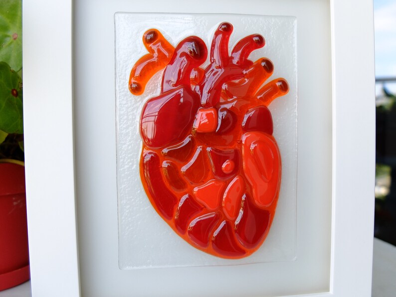 May include: A framed glass mosaic artwork depicting a red heart. The heart is made of many small pieces of red and orange glass.