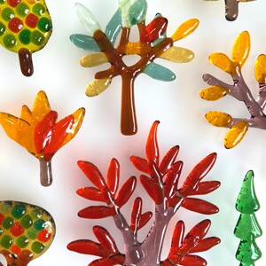 May include: A collection of colorful glass tree ornaments in various shapes and sizes. The trees feature shades of red, orange, yellow, green, and brown. Some have rounded leaves, while others have more pointed shapes, all against a white background.