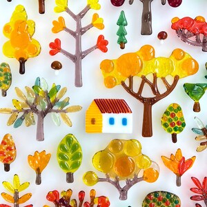May include: A collection of colorful glass tree and house figurines in shades of yellow, orange, green, and brown. The miniature trees vary in style, with some featuring rounded canopies and others with more detailed branch structures. A small house with a yellow body and red roof is also present.