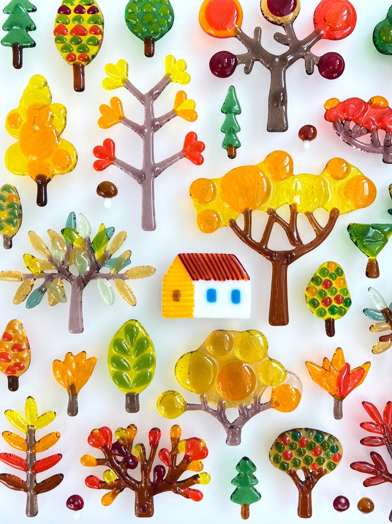 May include: A collection of colorful glass tree and house figurines in shades of yellow, orange, green, and brown. The miniature trees vary in style, with some featuring rounded canopies and others with more detailed branch structures. A small house with a yellow body and red roof is also present.