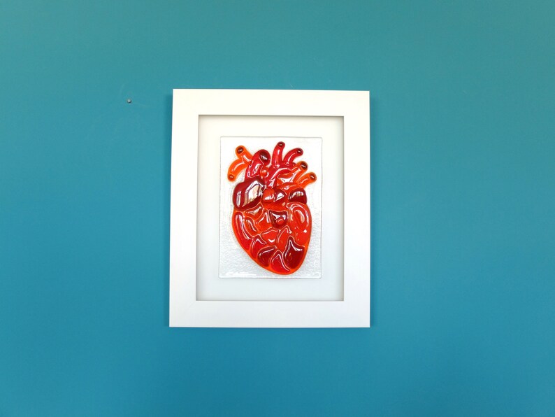 May include: A framed glass mosaic artwork depicting a red heart. The heart is made of many small pieces of glass in shades of red and orange.