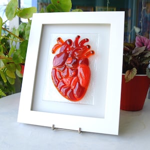 May include: A framed glass art piece depicting a red heart. The heart is made of many small pieces of red glass, giving it a textured appearance.
