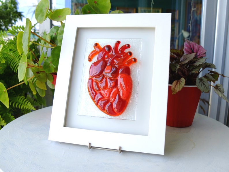 May include: A framed glass art piece depicting a red heart. The heart is made of many small pieces of red glass, giving it a textured appearance.