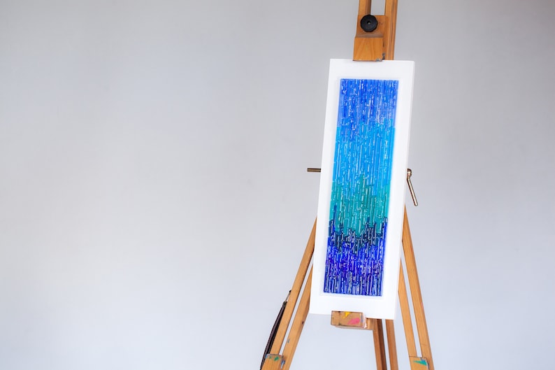 May include: Abstract art piece featuring a vertical composition of blue and teal hues. The artwork, mounted on a white frame, is displayed on a wooden easel. The color palette ranges from deep indigo to light turquoise, creating a gradient effect.