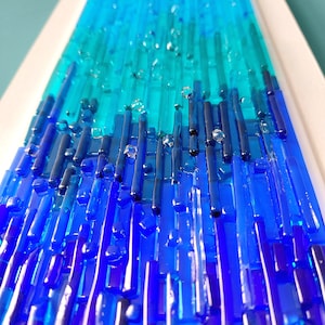 May include: Abstract art piece featuring a gradient of blues and teals. Vertical glass rods create a textured surface, transitioning from deep blue at the base to lighter shades at the top. Clear glass accents add dimension.