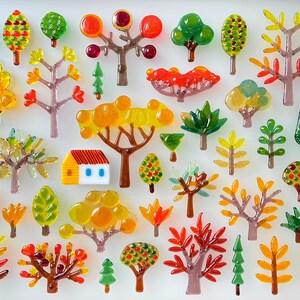 May include: A collection of colorful glass tree ornaments in various shapes and sizes. The trees feature shades of green, yellow, orange, and red, with some resembling autumn foliage. A small house ornament with a red roof and blue windows is also present.