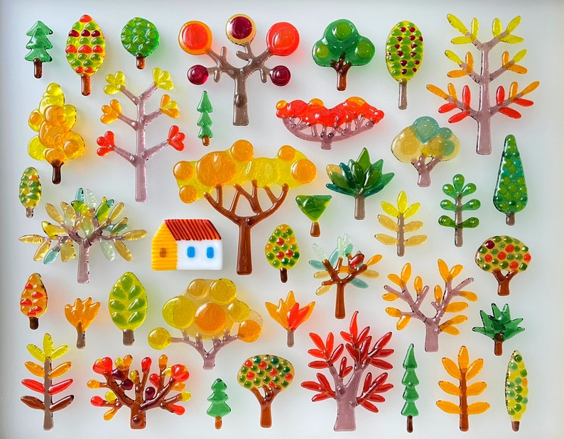 May include: A collection of colorful glass tree ornaments in various shapes and sizes. The trees feature shades of green, yellow, orange, and red, with some resembling autumn foliage. A small house ornament with a red roof and blue windows is also present.