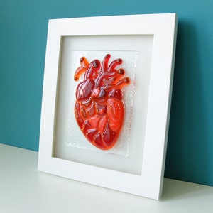 May include: A framed glass art piece depicting a red anatomical heart. The heart is made of stained glass and is set against a white background.