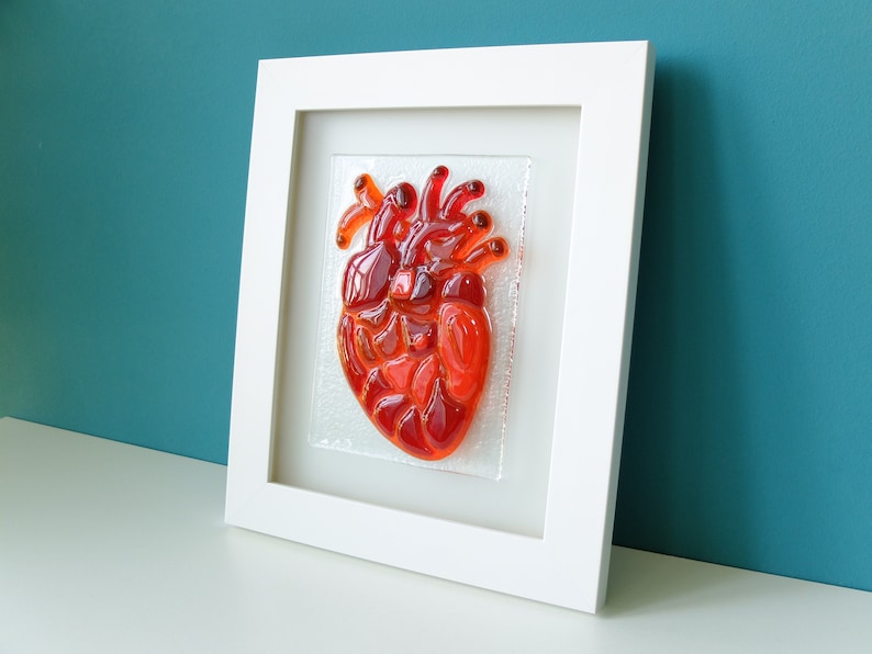 May include: A framed glass art piece depicting a red anatomical heart. The heart is made of stained glass and is set against a white background.