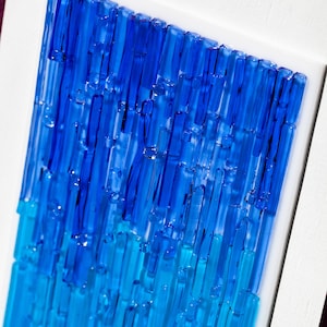 May include: Abstract art piece featuring a gradient of blue glass rods. The rods are arranged vertically, transitioning from a deep blue at the top to a lighter turquoise at the bottom. The artwork is set within a white frame.