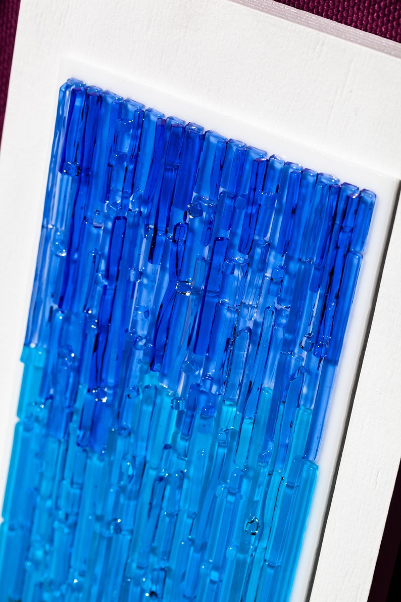 May include: Abstract art piece featuring a gradient of blue glass rods. The rods are arranged vertically, transitioning from a deep blue at the top to a lighter turquoise at the bottom. The artwork is set within a white frame.