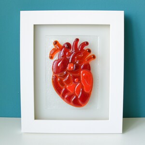 May include: A framed glass art piece depicting a red anatomical heart. The heart is made of many small pieces of red glass, creating a textured and dimensional effect.