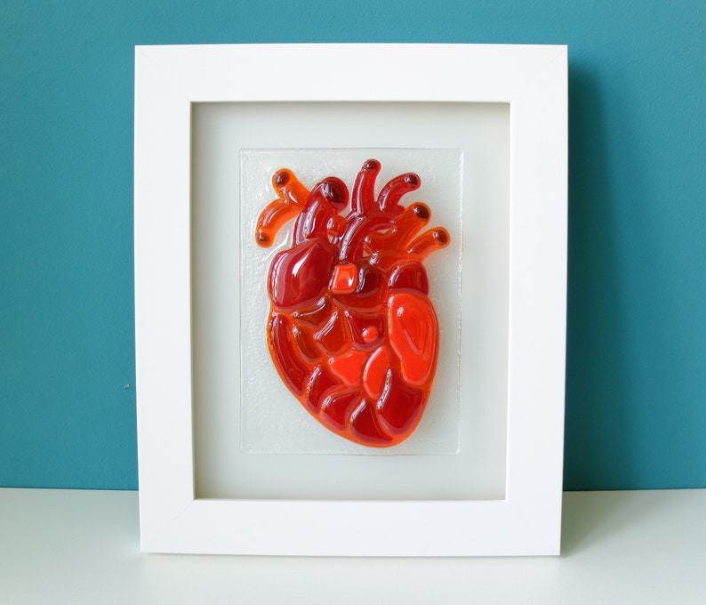May include: A framed glass art piece depicting a red anatomical heart. The heart is made of many small pieces of red glass, creating a textured and dimensional effect.