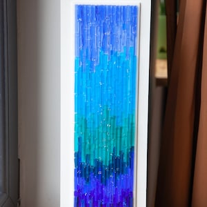 May include: A rectangular wall art piece featuring a gradient of blue and teal glass rods set in a white frame. The artwork transitions from deep indigo at the bottom to lighter shades of blue at the top, creating a visual flow.