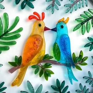 May include: Two glass birds perched on a branch, surrounded by green leaves. The bird on the left is yellow with orange accents, while the bird on the right is blue with green accents.