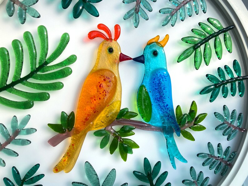 May include: Two glass birds perched on a branch, surrounded by green leaves. The bird on the left is yellow with orange accents, while the bird on the right is blue with green accents.