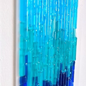 May include: Abstract glass art piece with a gradient of blues and turquoise. The artwork features vertical, rectangular glass pieces arranged in a layered pattern, creating a textured, three-dimensional effect. The piece is mounted on a white background.
