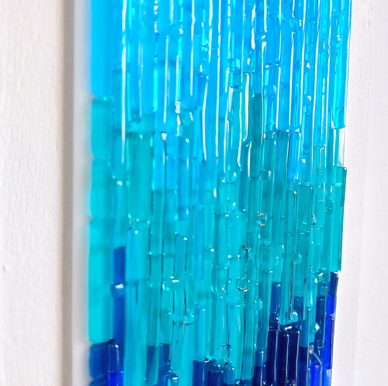 May include: Abstract glass art piece with a gradient of blues and turquoise. The artwork features vertical, rectangular glass pieces arranged in a layered pattern, creating a textured, three-dimensional effect. The piece is mounted on a white background.