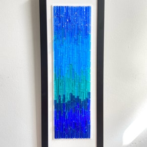 May include: A framed abstract art piece featuring a gradient of blue glass rods. The artwork transitions from dark blue at the bottom to lighter shades of blue and turquoise at the top, encased in a black frame.