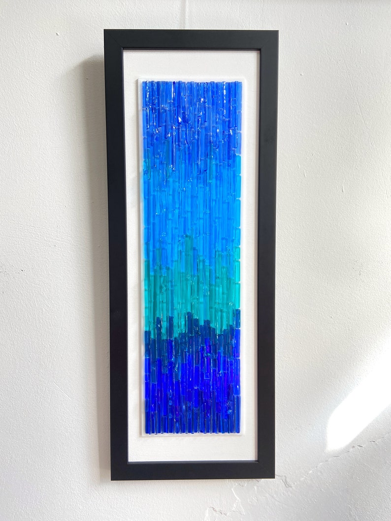 May include: A framed abstract art piece featuring a gradient of blue glass rods. The artwork transitions from dark blue at the bottom to lighter shades of blue and turquoise at the top, encased in a black frame.