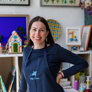 May include: A woman wearing a dark blue shirt and a dark blue apron with the text "Olga Glass" embroidered on the front. She is smiling and has her hand on her hip.