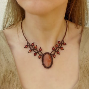 Puede incluir: Un collar de cordón marrón con un colgante de piedra de cornalina de forma ovalada. El collar presenta un diseño delicado de pequeñas cuentas rojas dispuestas en forma de hojas.