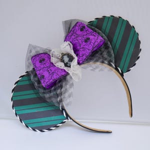 May include: A decorative headband with two circular ear shapes. The ears feature green and black stripes and are trimmed with black and white cording. A purple bow with a black spider web pattern and a cameo pendant adorns the center.