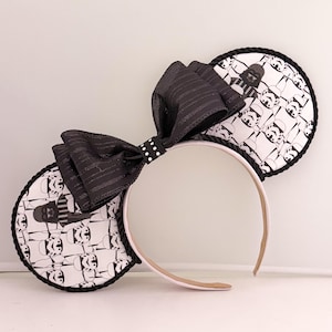 May include: A headband featuring two circular ears with a white fabric base printed with black and white illustrations of Darth Vader and Stormtroopers. The ears are trimmed with black twisted cord and a large black fabric bow sits in the center.