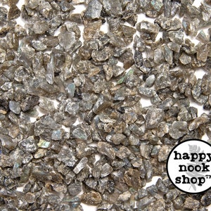 Könnte beinhalten: Nahaufnahme eines Haufens kleiner, irisierender Labradorit-Splitter. Die Splitter sind in verschiedenen Grautönen gehalten und haben eine schimmernde, reflektierende Qualität. Die Splitter sind auf einem weißen Hintergrund verstreut.