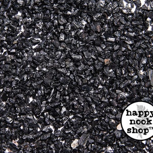 May include: A close-up image of black tourmaline crystals, also known as schorl, scattered on a white surface. The crystals are small and jagged, with a dark, opaque appearance. The image is labeled "happy nook shop" in the lower right corner.