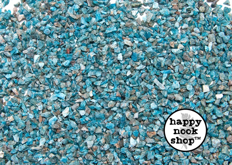 May include: A close-up view of a large quantity of small, blue-green, irregularly shaped, and translucent gemstone chips. The chips are scattered across a white surface. The image includes a small, round, white logo with the text "happy nook shop" in black.