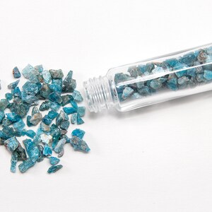 May include: A small glass bottle filled with blue and green gemstone chips. Some of the chips have spilled out of the bottle onto a white surface.