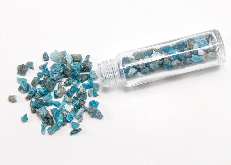 May include: A small glass bottle filled with blue and green gemstone chips. Some of the chips have spilled out of the bottle onto a white surface.