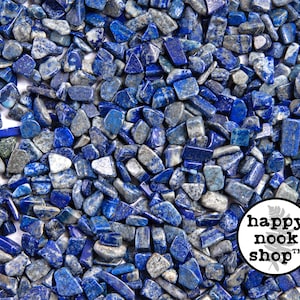 May include: A close-up image of a large quantity of small, irregularly shaped, blue and grey lapis lazuli stones. The stones are scattered on a white surface. The image includes the text "happy nook shop" in a circle with a leaf design.