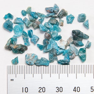 May include: A collection of blue-green gemstone chips, some with a darker green hue, scattered on a white surface. A ruler is placed below the chips, with markings in millimeters from 0 to 60.