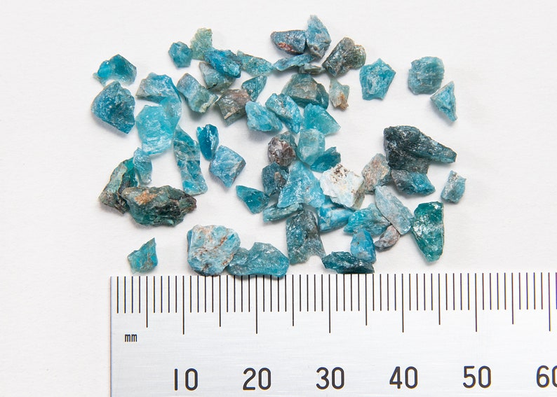 May include: A collection of blue-green gemstone chips, some with a darker green hue, scattered on a white surface. A ruler is placed below the chips, with markings in millimeters from 0 to 60.