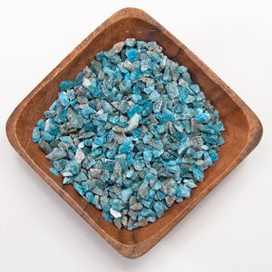 May include: A wooden bowl filled with blue apatite crystal chips. The chips are a light blue color with some darker blue and gray accents.