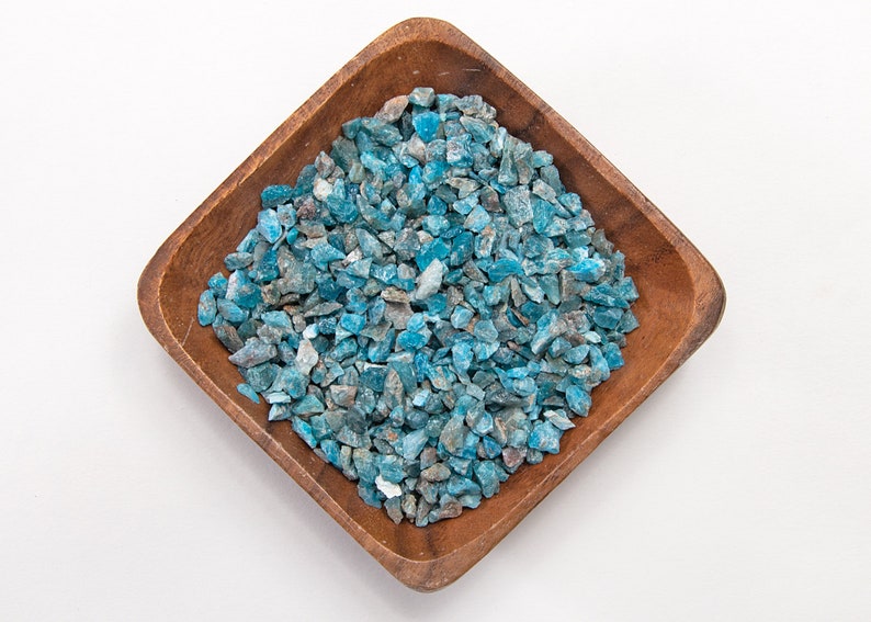May include: A wooden bowl filled with blue apatite crystal chips. The chips are a light blue color with some darker blue and gray accents.