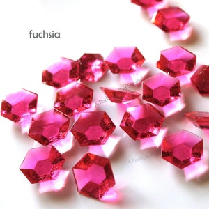 May include: A scattering of fuchsia-colored, faceted, hexagonal-shaped, clear plastic gems. The gems are scattered on a white surface.