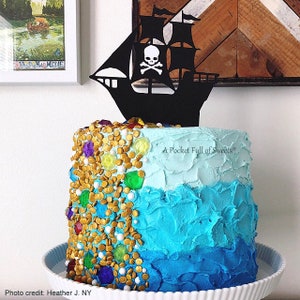 Peut inclure: Un g&acirc;teau ombr&eacute; bleu et blanc d&eacute;cor&eacute; de p&eacute;pites dor&eacute;es et de couleurs de pierres pr&eacute;cieuses. Une silhouette noire d'un navire pirate avec un cr&acirc;ne et des os crois&eacute;s est plac&eacute;e sur le dessus du g&acirc;teau. Le g&acirc;teau est sur un support &agrave; g&acirc;teau blanc. Le texte "A Pocket Full of Sweets" est sur le c&ocirc;t&eacute; du g&acirc;teau.